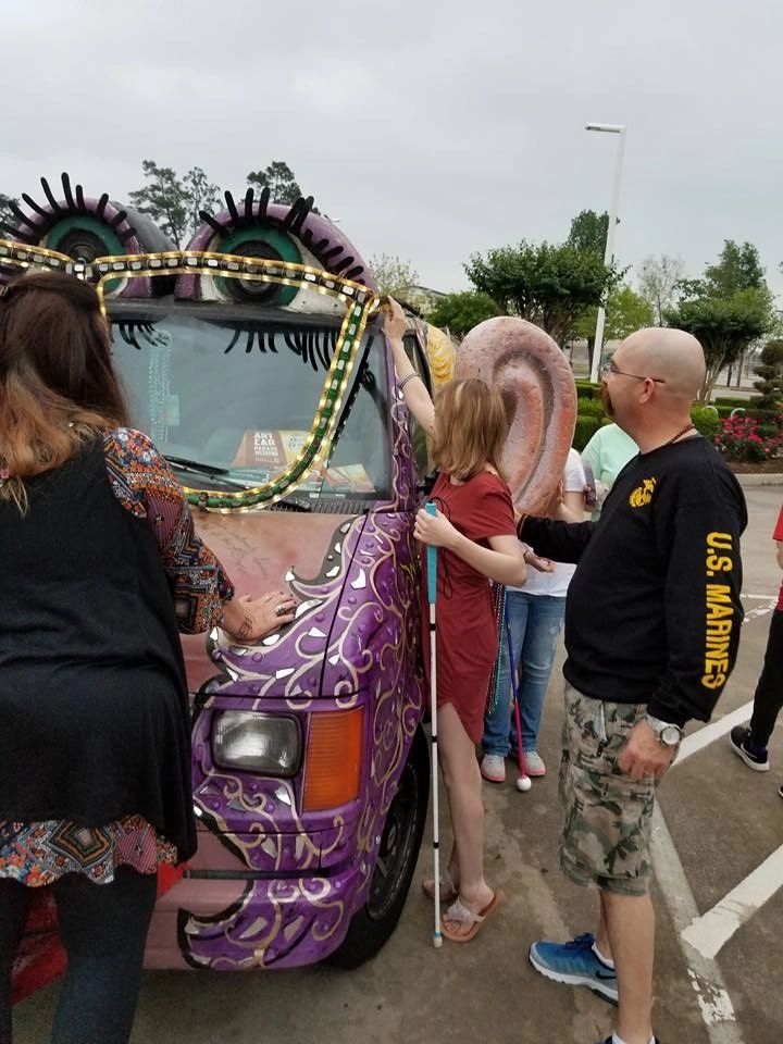 An artist reaches up to touch the sculpted ear on the Women Rock Art Car while a volunteer stands close beside them, a white cane and beaded strands visible nearby.