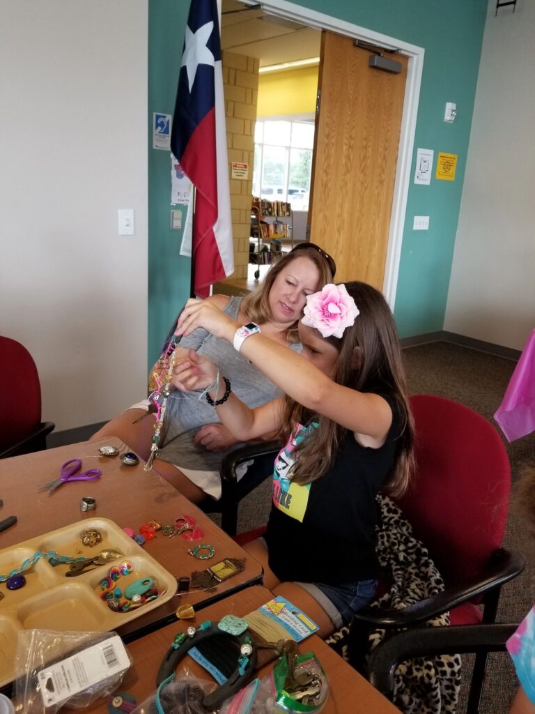 A volunteer helps an artist attach a beaded strand to a horseshoe necklace, more beads and charms spread on the table.