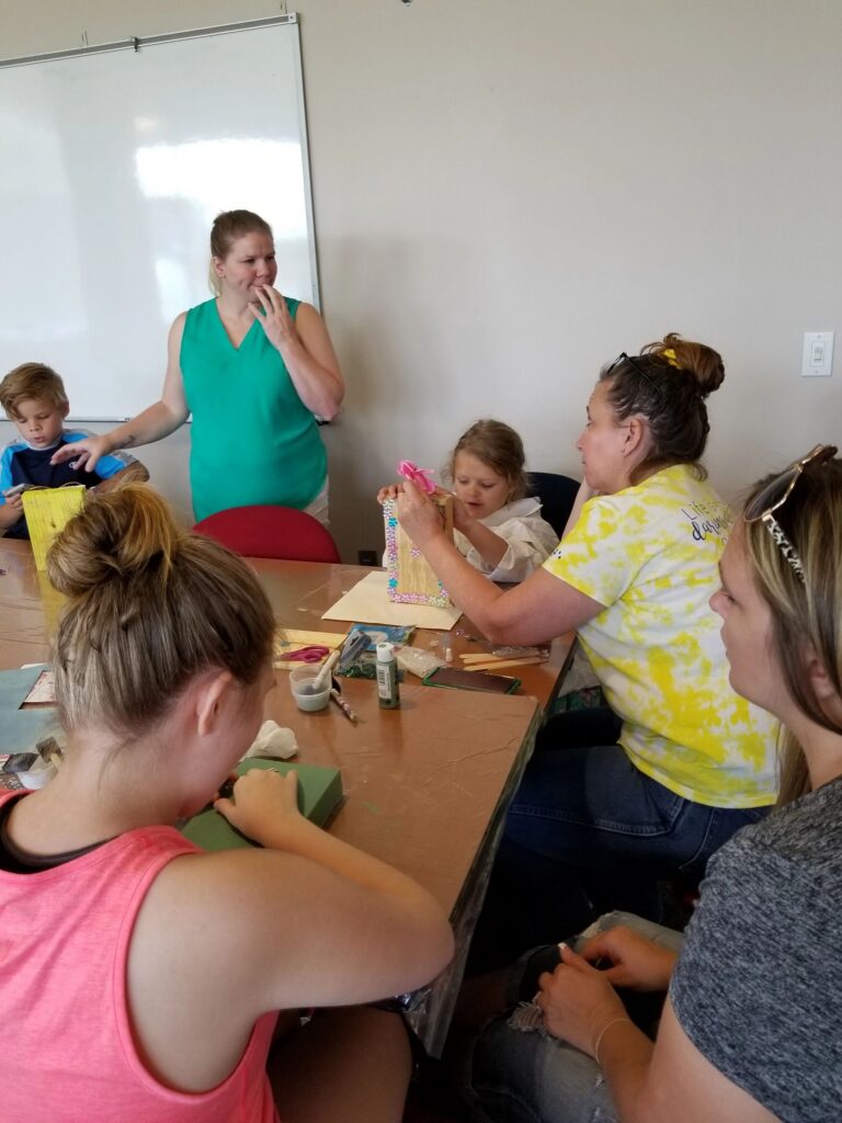 Artists sit around a table decorating wooden boxes, one holding a pink ribbon flower, craft supplies spread across the tabletop.