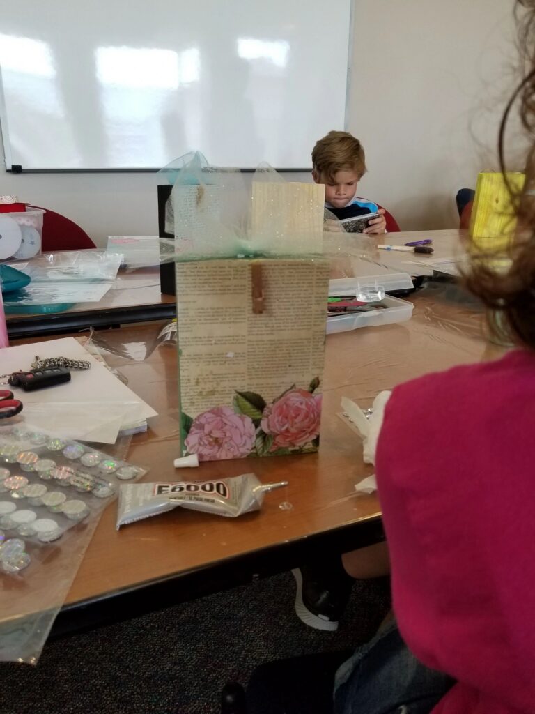 A wooden box wrapped in vintage rose-print paper with a decorative clothespin accent, tissue paper draped over the top, resting on a craft table.