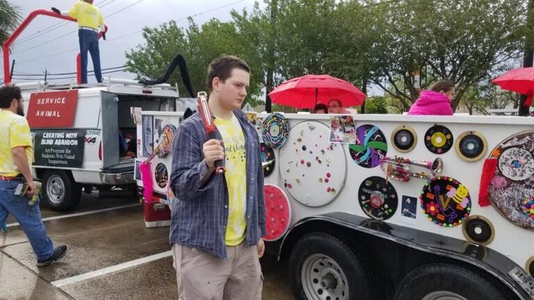 A rider holds a colourful glass bead braille stick, standing near the float's decorated panel of records, a trumpet and painted circles.