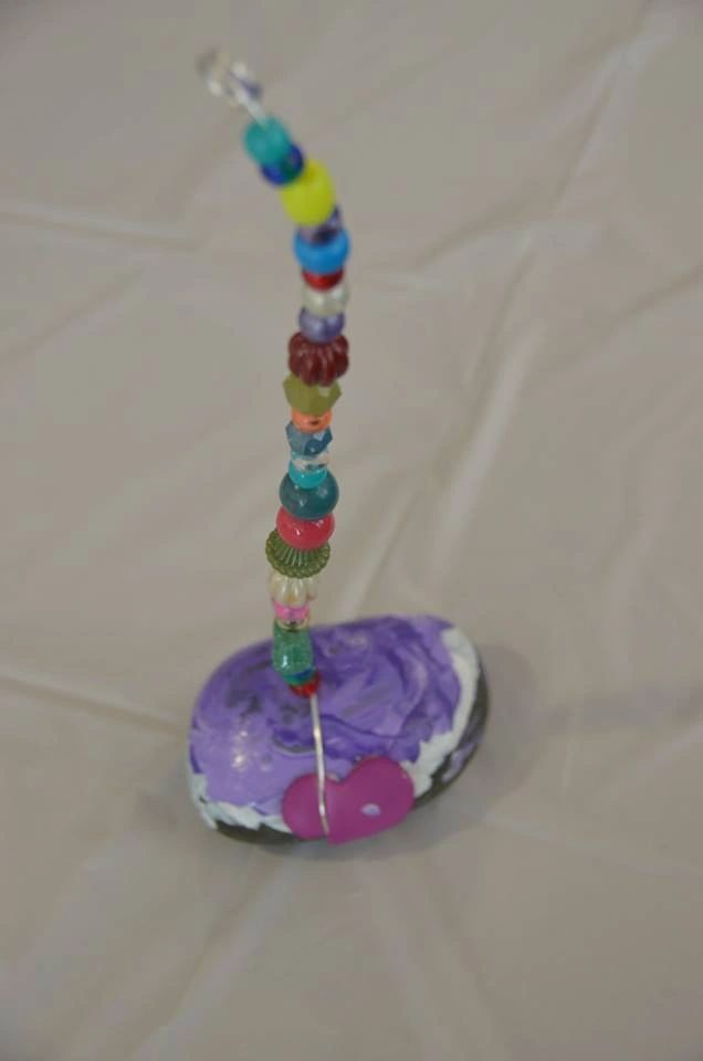 A beaded wand rising from a purple-painted stone base decorated with a small pink heart charm, the beads in shades of blue, red, yellow and green.