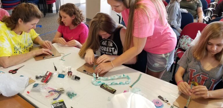 Artists and a volunteer work together beading on clipboards, nail polish and braided cord spread across the table.