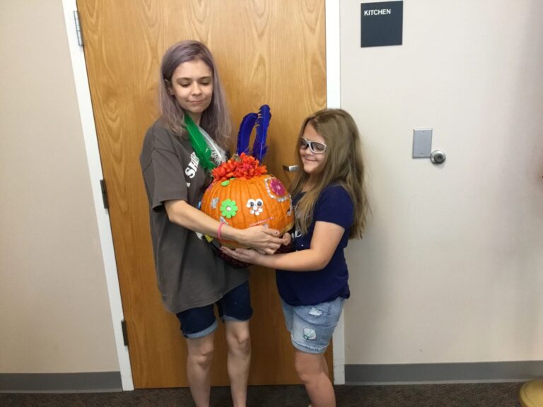Two artists hold up a pumpkin covered in painted flowers and a face made from googly eyes, topped with green and blue feathers.