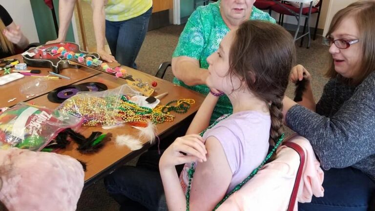 Artists and volunteers sit around a table covered in beads, ribbon and decorated records, one artist having beaded strands placed in their braided hair.