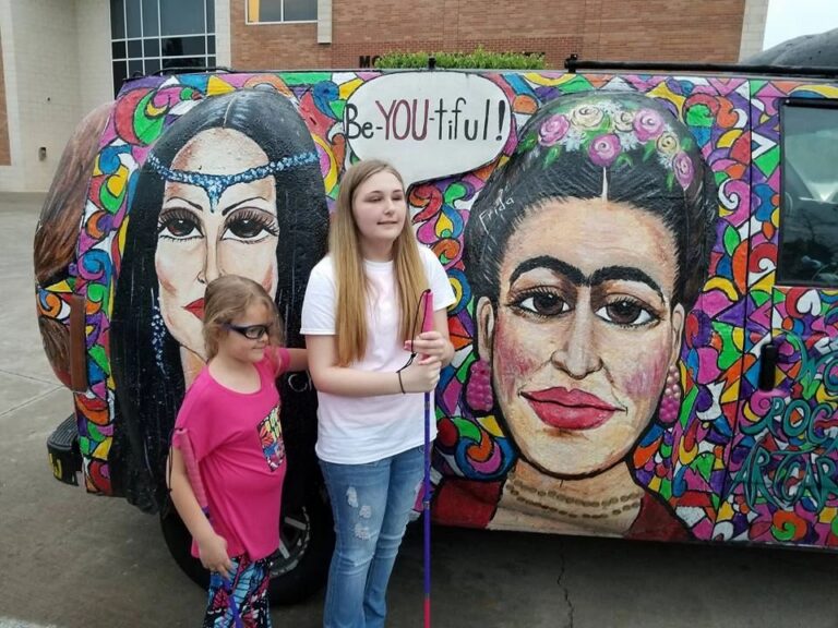 Two artists stand before the Women Rock Art Car, painted with portraits of famous women and a Be-YOU-tiful speech bubble, one holding a white cane.