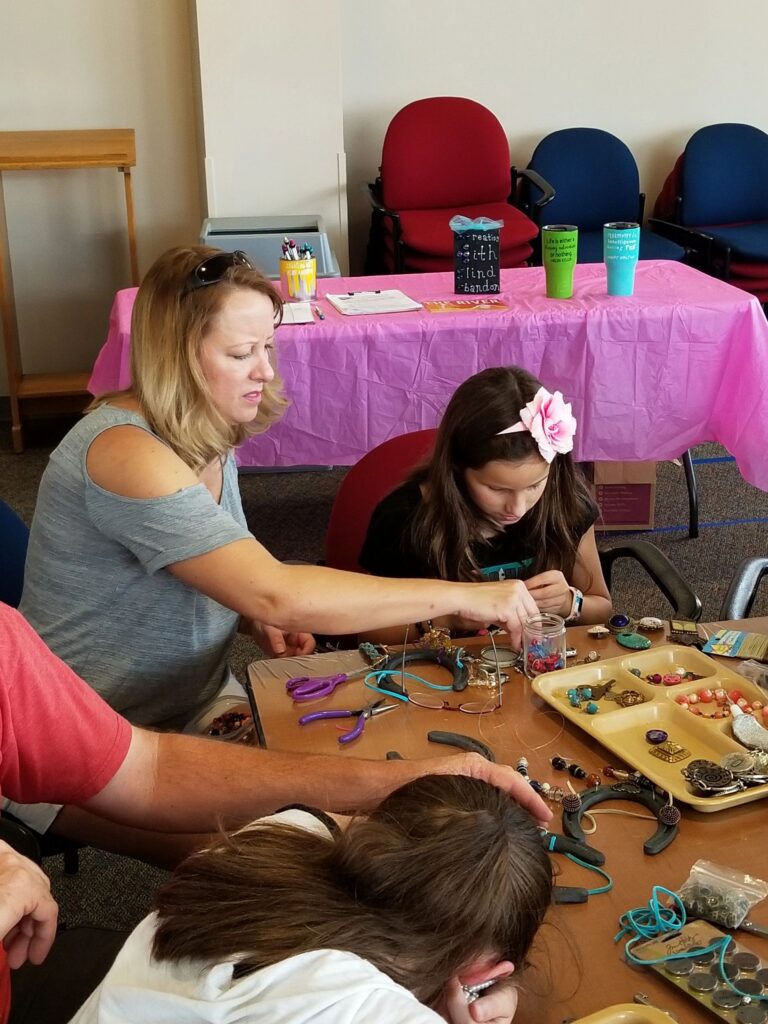 A volunteer helps an artist thread beads onto a horseshoe pendant, trays of charms and small trinkets on the table.