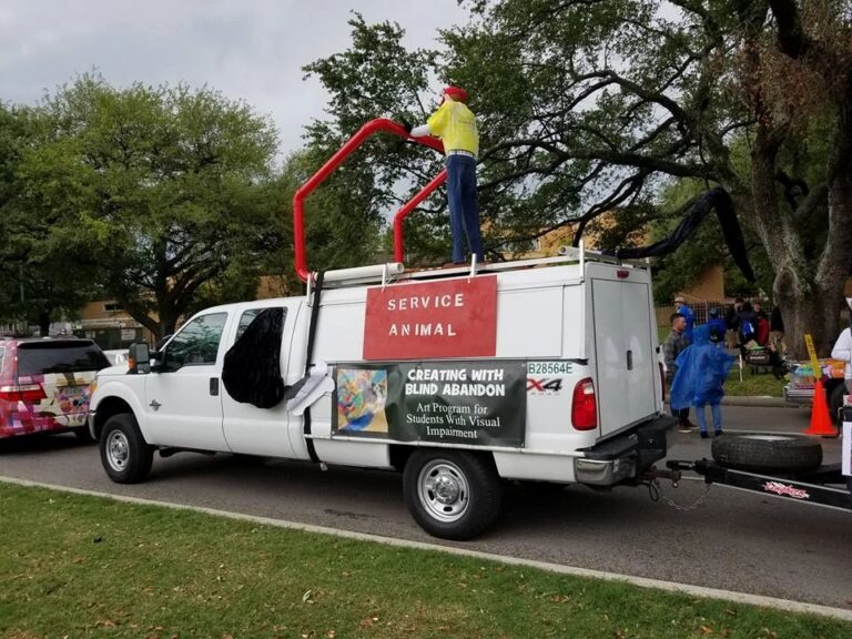 A pickup truck towing the float, with a rider standing on a raised platform on the truck's roof and a Service Animal sign and program banner on the truck bed.
