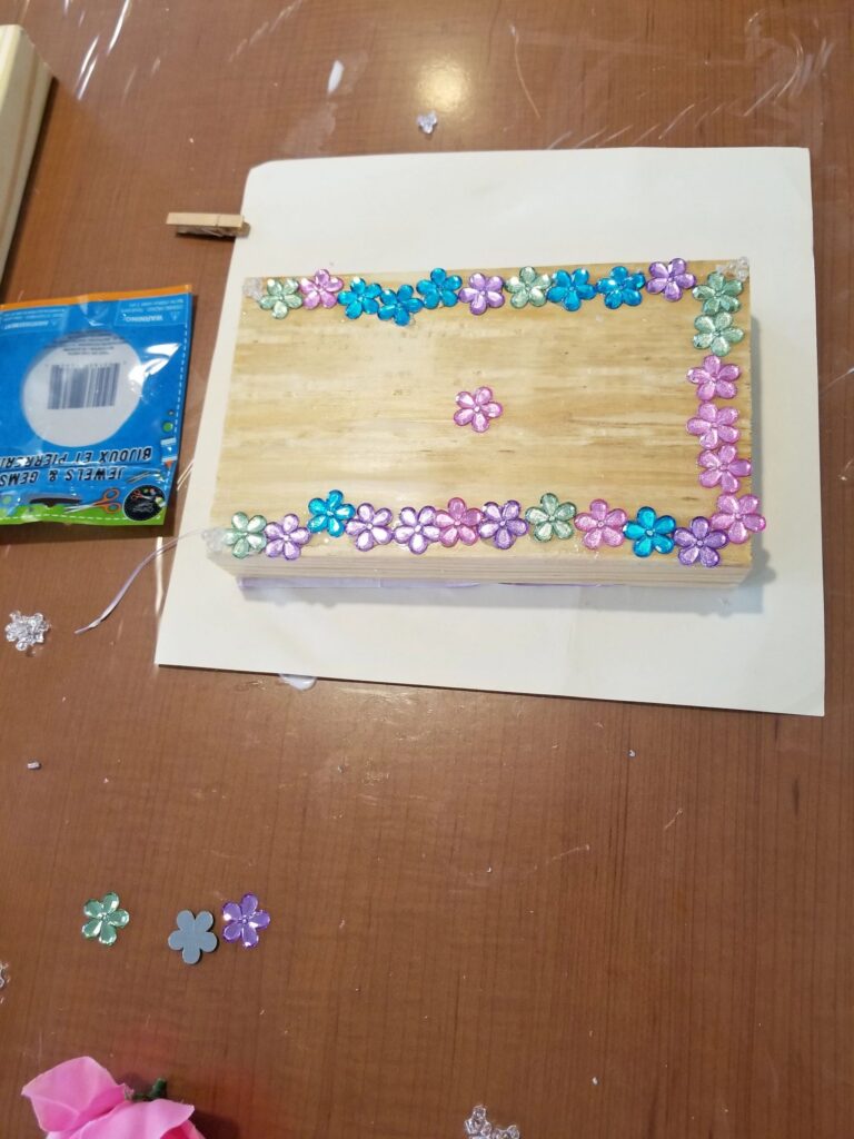 A small wooden frame bordered with sparkling flower gems, a single pink gem in the centre, resting on a sheet of paper.