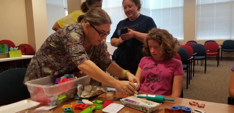 A volunteer helps an artist paint a dough ornament, cookie cutters and trays of shaped pieces spread across the table.