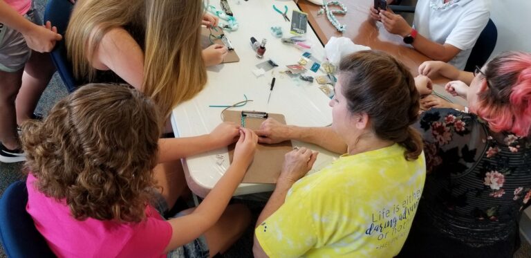 A volunteer helps an artist bead on a clipboard while other artists work around the table, beads and small tools scattered nearby.