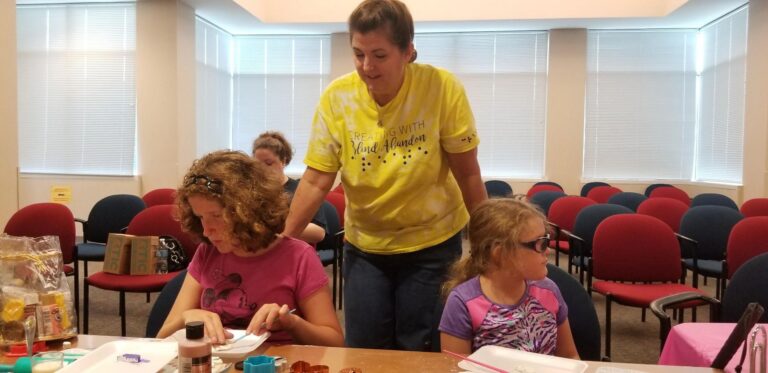 A volunteer helps two artists cut dough shapes with cookie cutters, trays of shaped ornaments visible on the table.