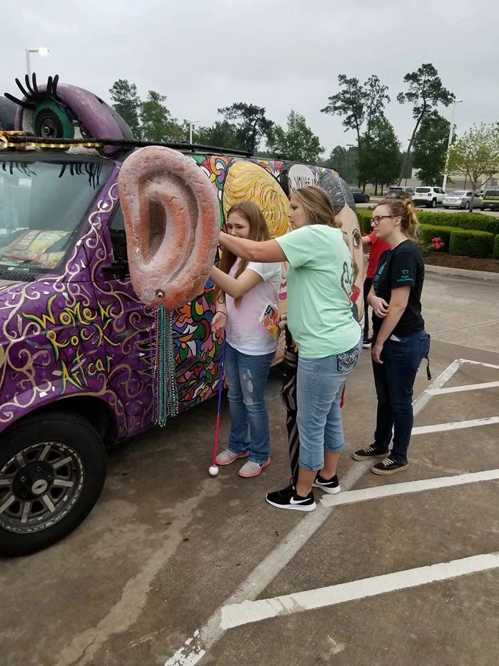 Several people reach out to touch a large sculpted ear mounted on the side of the Women Rock Art Car, painted with lash-lined eyes above.