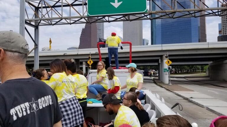 Three people in matching yellow tie-dye Creating With Blind Abandon shirts pose in front of the float, the giant stuffed hound visible behind them.
