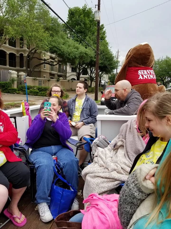 A group of parade-goers stands together on the sidewalk near the float, one person's arm wrapped comfortingly around another.