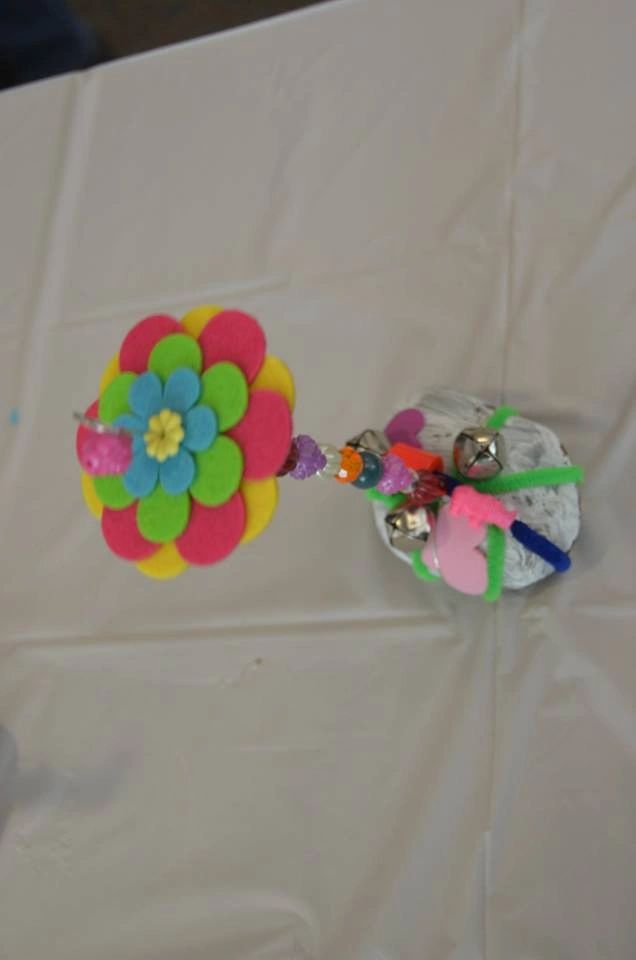 A painted stone decorated with a felt flower in pink, green and blue layers, joined by a beaded stem hung with small silver bells.