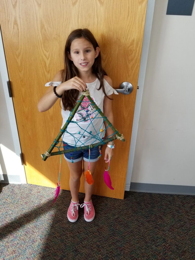 An artist holds up a finished triangular dream catcher woven with coloured yarn and hung with feathers on ribbon.