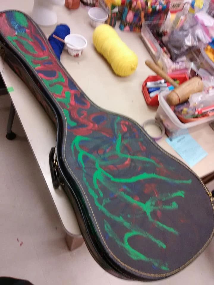 A guitar case painted with swirling green vines over a red and blue background, resting on a supply table with yarn, cups and craft bins nearby.