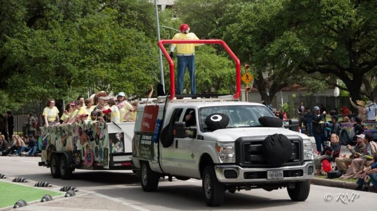 The truck towing the float drives down the parade route, riders waving from the platform on its roof, spectators lining the street on both sides.