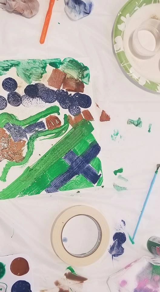 A close-up of a painted canvas with paint dabbed in green, blue, brown and black shapes, brushes and a paint palette resting nearby.