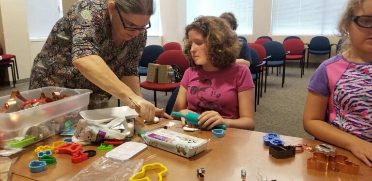 A volunteer arranges cookie cutters shaped as letters, animals and stars on a table, artists seated nearby with baby wipes and small tools.