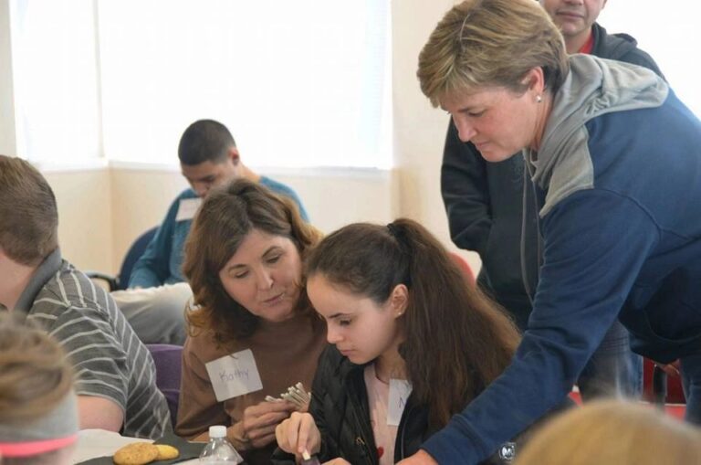 A volunteer and an artist look closely at a beaded craft piece together at a table set with cookies and water.