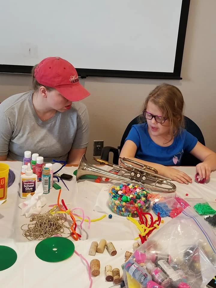 A volunteer and an artist decorate a trumpet with beads and pipe cleaners at a table covered with corks, bottle caps and craft supplies.