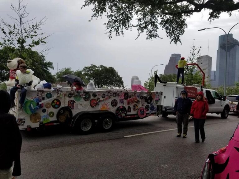The float travels down the parade route past downtown high-rises, riders under an umbrella and a decorated record visible on the panel.