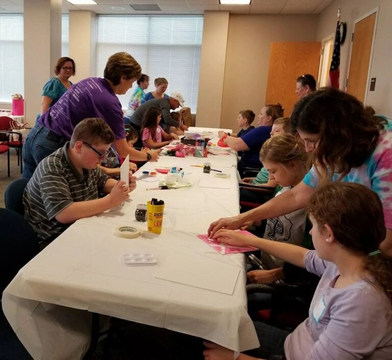A wide view of the studio, with artists and volunteers seated around a long table working on projects with paint trays and craft supplies spread out.