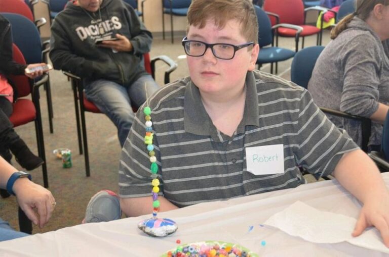 An artist sits with a beaded wand made from a painted stone base and a colourful strand of beads rising from it, seated among rows of chairs.