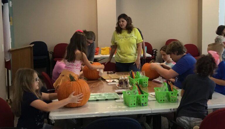 A volunteer and artists carry and decorate pumpkins at a table set with paint trays and supply baskets.