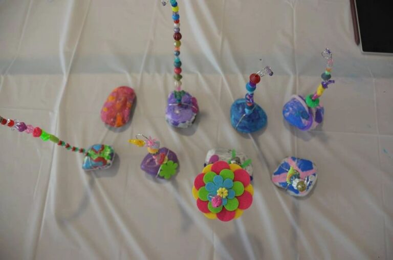 A group of painted stones and beaded wands displayed together: some stones painted with swirled patterns, others with simple flower shapes, each topped with a colourful beaded stem.