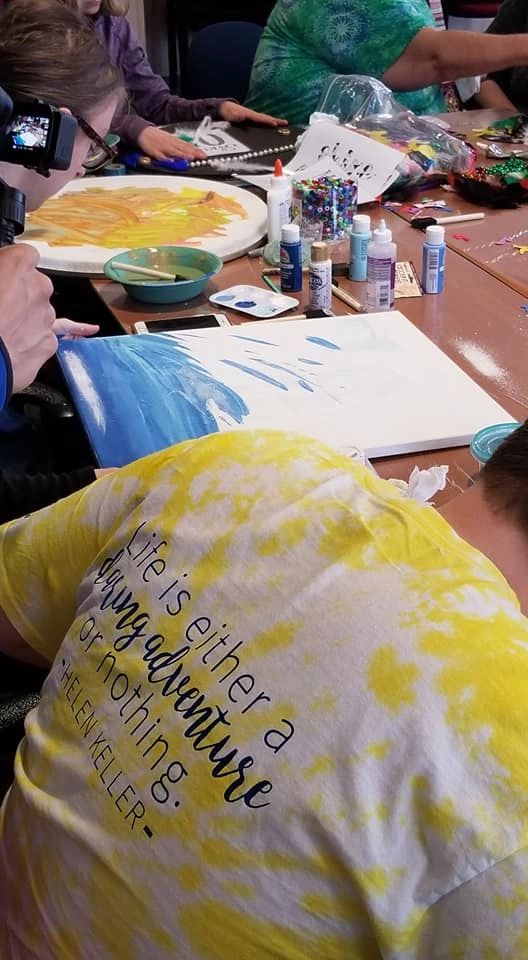 An artist paints a blue canvas at a table covered in craft supplies, sitting beside someone wearing a yellow tie-dye shirt printed with a Helen Keller quote.