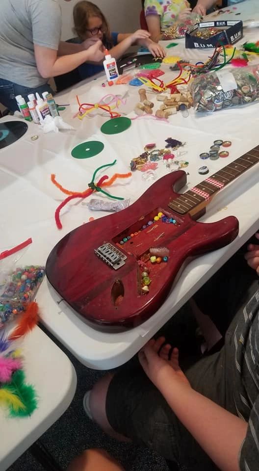 An electric guitar body decorated with a cluster of colourful beads set into the pickup cavity, resting on a table covered in craft supplies.