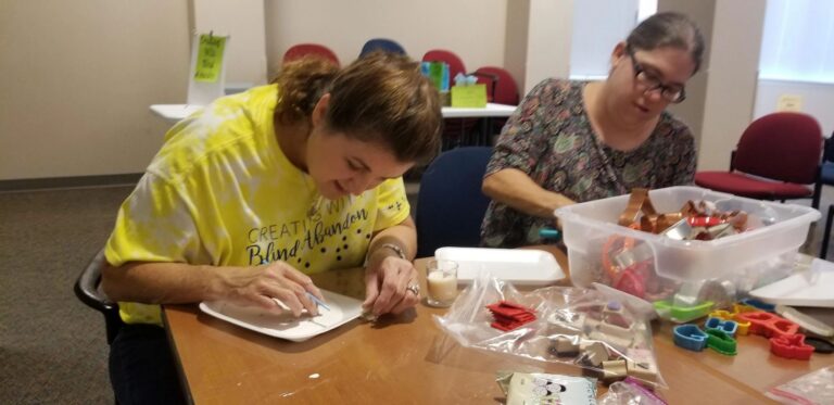 A volunteer helps an artist paint a dough shape, more cookie cutters and shaped pieces spread across the table.