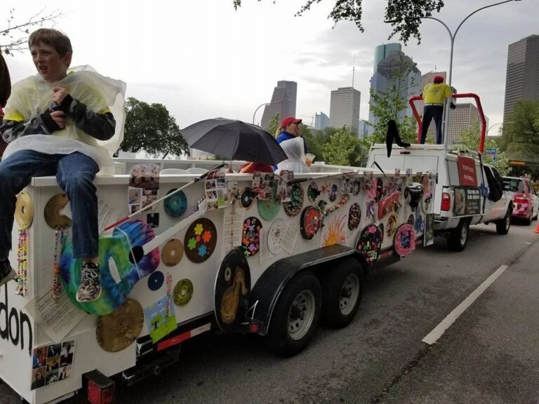 A rider sits on the edge of the float, decorated records and a tie-dye guitar visible on the panel beside them, downtown high-rises in the background.