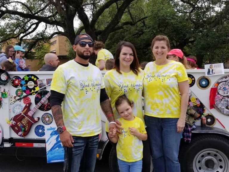Four people in matching yellow Creating With Blind Abandon t-shirts pose together in front of the parade float, one shirt printed with raised braille dots.