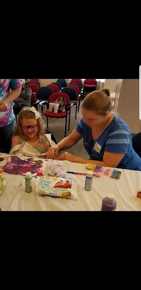 A volunteer helps an artist with a craft project at a table set with paint bottles and baby wipes, empty chairs visible behind them.