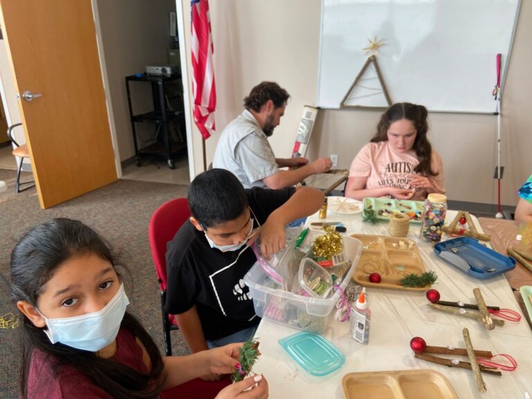 Artists around a worktable assembling stick Christmas trees, with trays of buttons, jingle bells, red ornaments and sprigs of greenery spread across the table. A finished twine-wrapped tree stands at the edge of the frame.