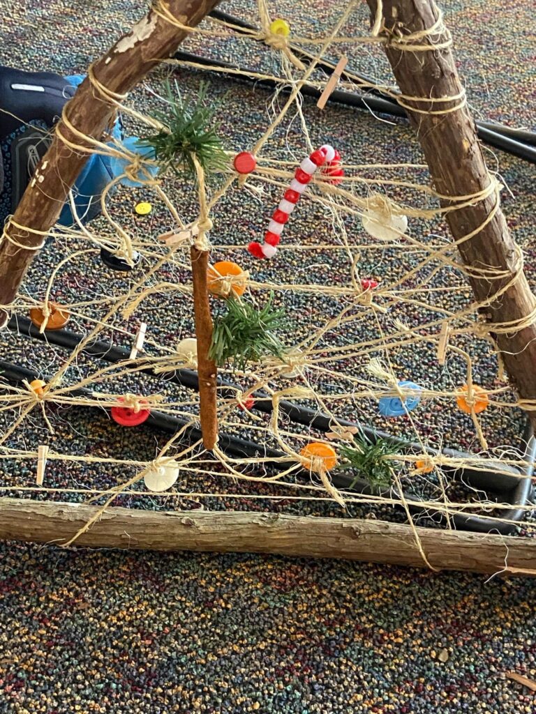 A finished stick-and-twine Christmas tree laid on the carpet. Twine is strung in horizontal rows across the triangular frame and hung with buttons, wooden clothes pegs, a beaded red and white candy cane, a cinnamon stick and small tufts of artificial pine.