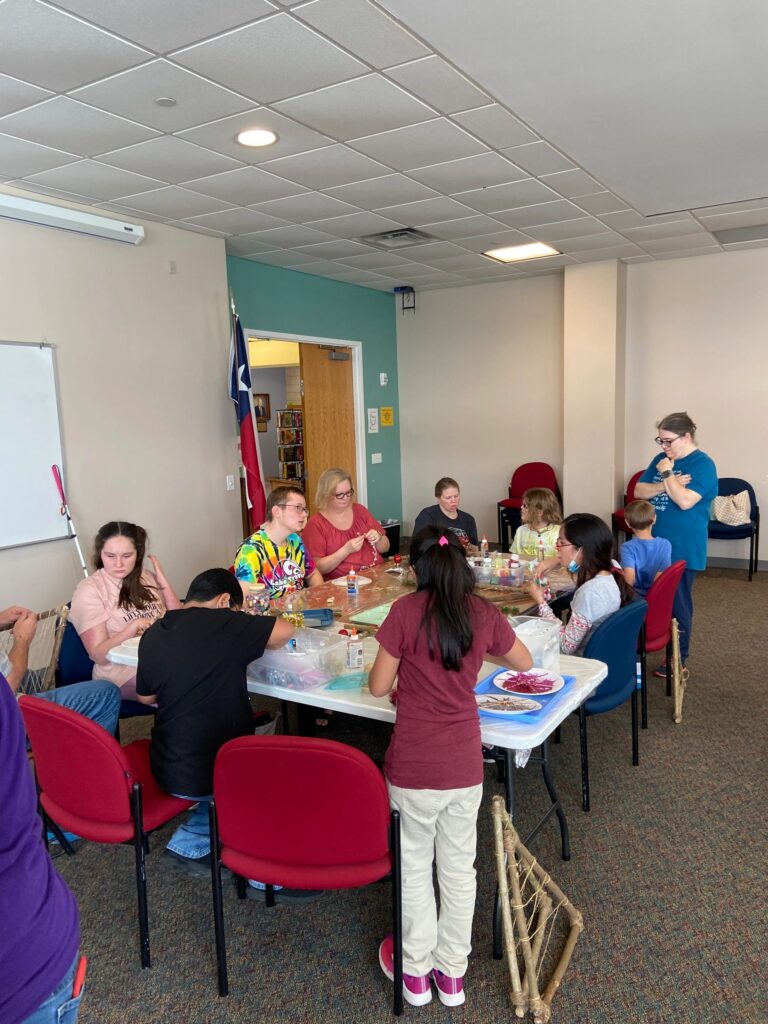 A wide view of the December session. Artists and volunteers work around a long table covered in supplies, with a completed stick tree resting against a chair in the foreground and a white cane leaning nearby.
