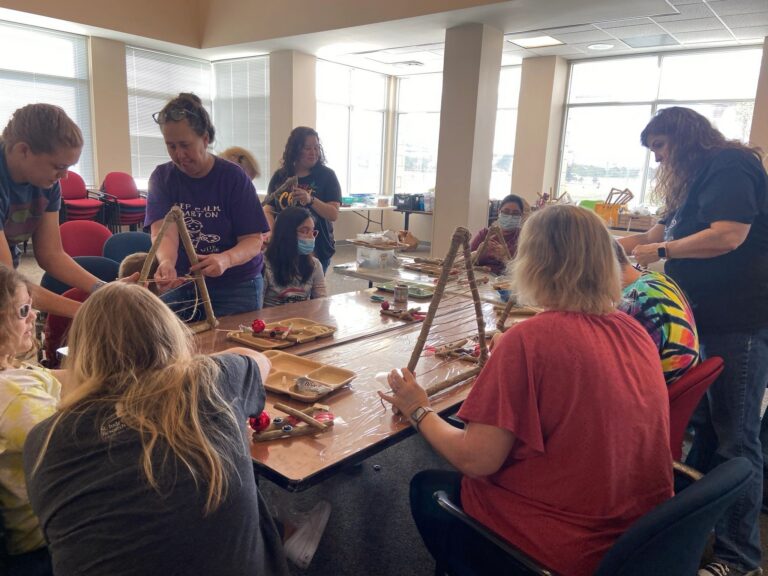 Artists and volunteers at a large table, several holding triangular stick frames as they wind twine between the branches. Trays of red ornaments and cinnamon sticks sit within easy reach.