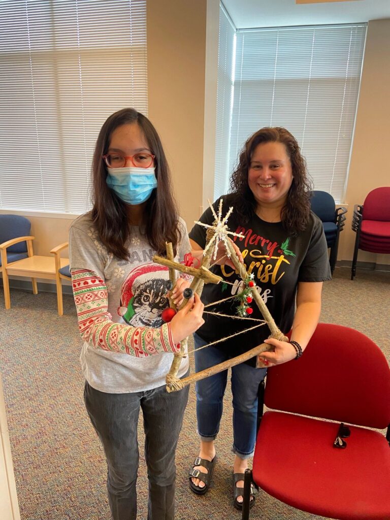 An artist and a volunteer hold up a completed stick Christmas tree together. The triangular branch frame is bound with twine at the corners, topped with a woven straw star and hung with red beads and greenery.