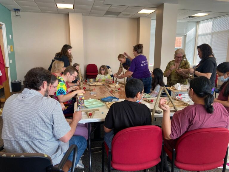 A busy worktable seen from one end, with artists and volunteers wrapping twine around branch frames. Jars of beads, red ornaments and wooden trays of materials cover the table.