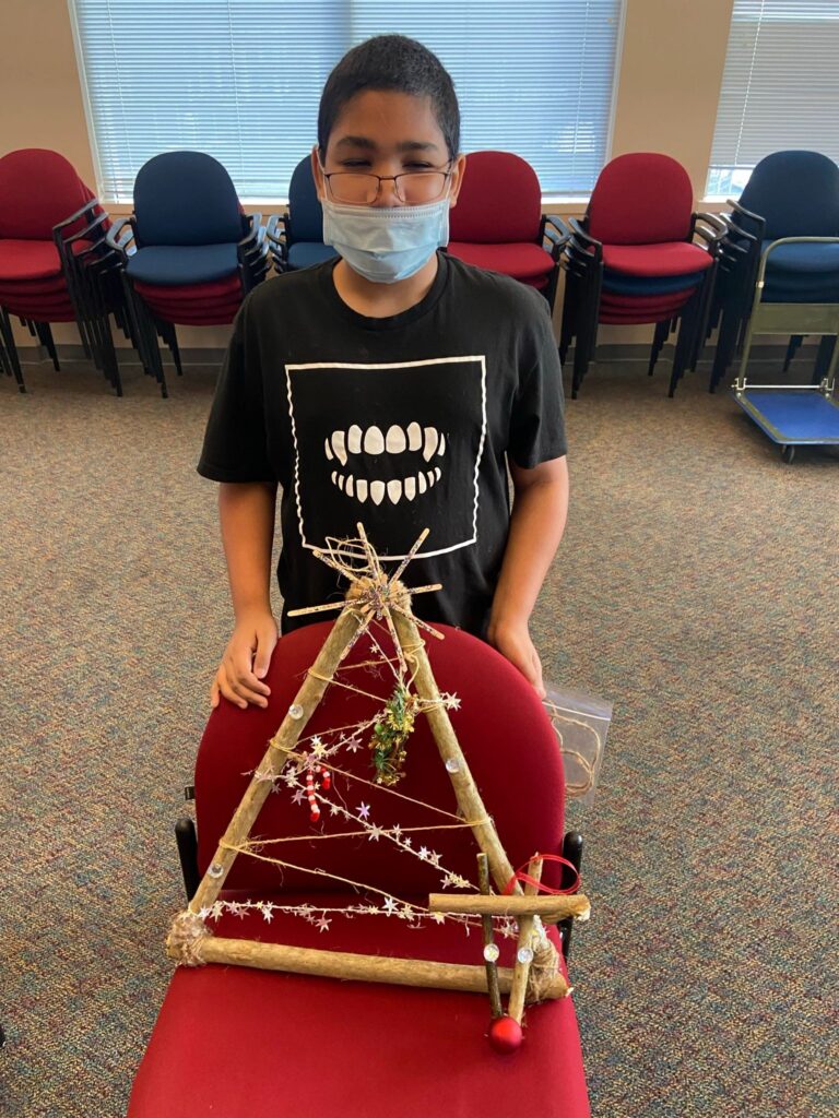 An artist stands behind a chair displaying a completed stick tree. The frame is criss-crossed with twine and trimmed with a silver star garland, a beaded candy cane and a cluster of artificial pine.