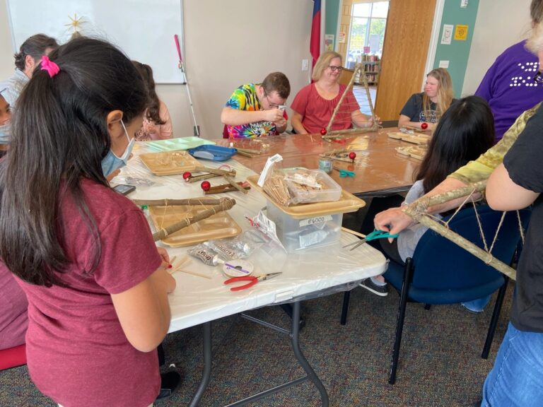 Artists working closely at a table, winding twine across branch frames. Trays of cinnamon sticks, craft sticks and red ornaments sit between them, with a whiteboard and a finished tree in the background.