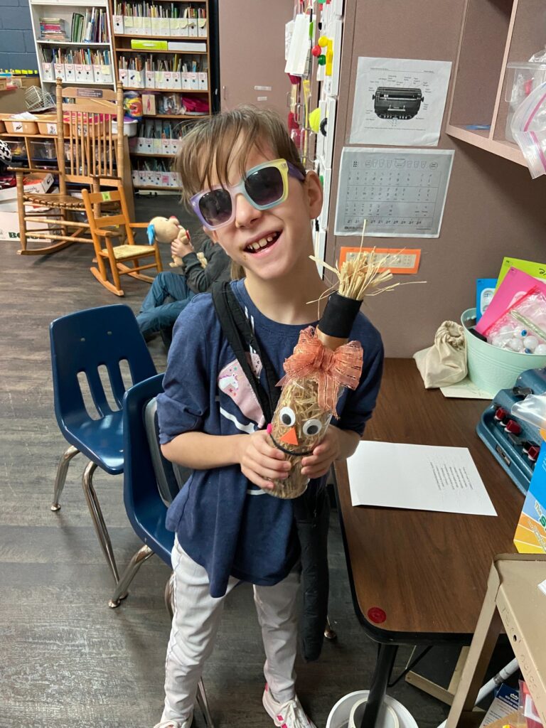 An artist holds a decorated cardboard tube figure with googly eyes, an orange paper beak and a raffia crown, finished with a wide organza bow.
