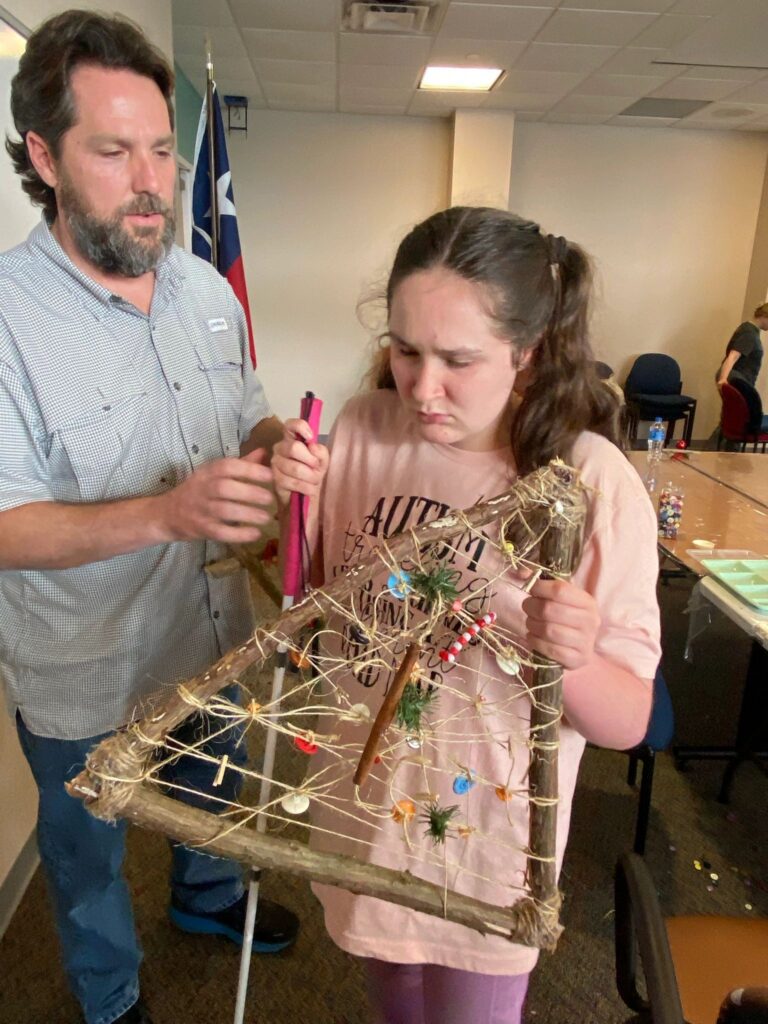A volunteer guides an artist's hands over a finished stick tree, helping trace the twine rows and the beads, cinnamon stick and greenery tied along them. The artist's white cane rests against them.