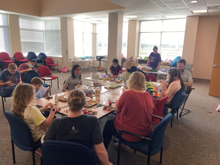 A wide view of the session room, with artists seated around two long tables working on their trees. Supplies, trays and finished pieces are spread across the tables.