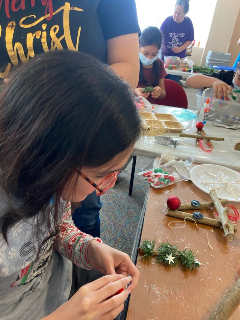 An artist threads a red bead onto twine, working close to the tabletop. Beside them lie branch pieces bound with twine, a red ornament, and sprigs of greenery with a white star.